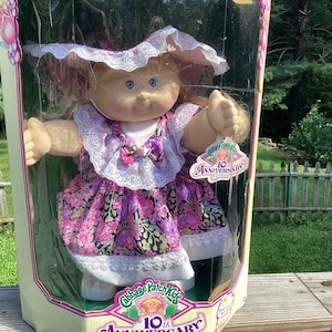 May include: A Cabbage Patch Kids 10th Anniversary doll with blonde yarn hair and blue eyes, wearing a purple and pink floral dress with white lace trim and a matching bonnet, encased in a clear plastic display box with anniversary branding.