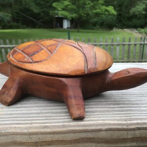 May include: A hand-carved wooden turtle box with a removable shell lid. The body is a dark brown wood, while the shell features a lighter tan finish with deep, incised geometric patterns. The turtle has four legs, a pointed tail, and a head.