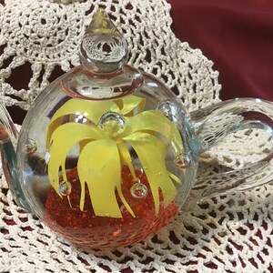 May include: A clear glass teapot with a yellow flower inside and red glitter at the bottom.