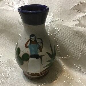May include: A small white ceramic vase with a blue rim and a painted design of a woman carrying a basket. The woman is wearing a blue shirt and a white skirt. The vase is decorated with green cactus plants.