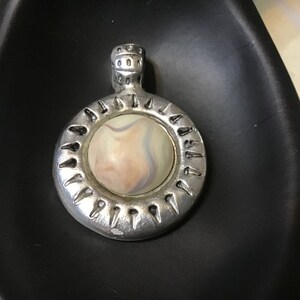 May include: A silver pendant with a round, white, pink, and blue swirl design in the center. The pendant has a decorative border with small points around the edge.