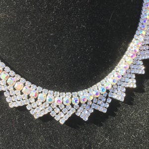 May include: A sparkling silver necklace with iridescent gemstones. The necklace features a series of clear, square-cut crystals and iridescent, oval-shaped stones. The design is set against a dark background, highlighting the necklace's brilliance.