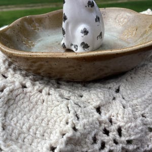 Puede incluir: Un plato de cer&aacute;mica hecho a mano con un exterior marr&oacute;n moteado y un interior azul claro. Una peque&ntilde;a figura blanca con manchas negras se encuentra dentro del plato. El plato descansa sobre un mantelito de ganchillo color crema.