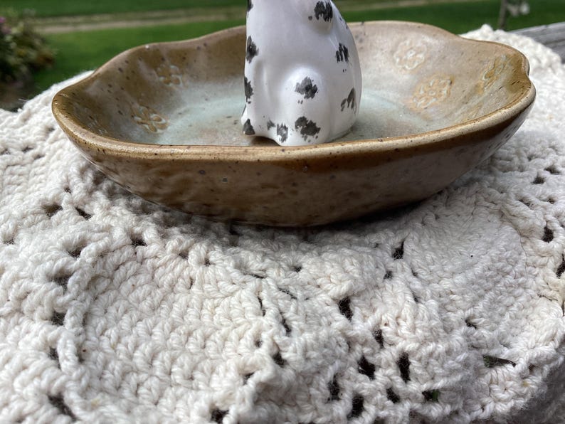 Puede incluir: Un plato de cer&aacute;mica hecho a mano con un exterior marr&oacute;n moteado y un interior azul claro. Una peque&ntilde;a figura blanca con manchas negras se encuentra dentro del plato. El plato descansa sobre un mantelito de ganchillo color crema.