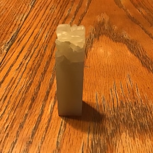 May include: A rectangular, light-colored stone seal with a textured top. The seal is standing upright on a wooden surface with a natural wood grain pattern. The seal's color is a pale, translucent hue.