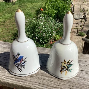 May include: Two white ceramic bells with gold accents. Each bell features a bird illustration with colorful details. The handles are elongated and topped with gold. The bells are displayed on a wooden surface.