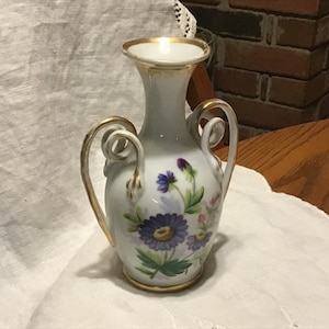 May include: A white porcelain vase with gold trim and floral designs. The vase has two handles and a narrow neck.