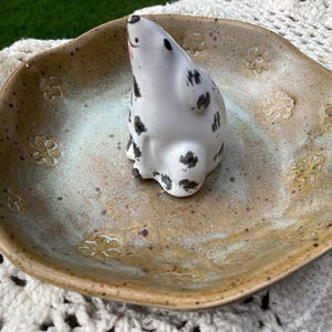 Puede incluir: Un plato de cer&aacute;mica con un esmalte marr&oacute;n claro moteado y grabados florales. Una figura de foca de cer&aacute;mica blanca con manchas negras se encuentra en el centro. El plato tiene un centro azul claro y blanco.