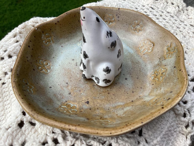 Puede incluir: Un plato de cer&aacute;mica con un esmalte marr&oacute;n claro moteado y grabados florales. Una figura de foca de cer&aacute;mica blanca con manchas negras se encuentra en el centro. El plato tiene un centro azul claro y blanco.