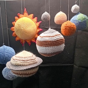 Pode incluir: Um móbile suspenso com um sol de crochê amarelo com raios laranja e várias esferas de crochê representando planetas em várias cores, incluindo azul, laranja, branco, marrom e cinza, suspensas por fios finos contra um fundo escuro.