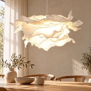 Op de afbeelding: Een witte hanglamp met een gelaagde, geplooide, papierachtige kap die een warm, diffuus licht verspreidt. De structuur bestaat uit meerdere overlappende, organisch gevormde lagen die aan een dunne, rechte metalen stang zijn opgehangen.