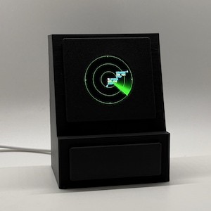 Könnte beinhalten: Ein schwarzes, angewinkeltes Desktop-Anzeigegerät mit einem kreisförmigen Bildschirm, der eine grüne Radarschnittstelle mit konzentrischen Ringen und digitalen Flugdaten zeigt. Das Gerät hat einen rechteckigen Sockel und ein weißes Stromkabel.