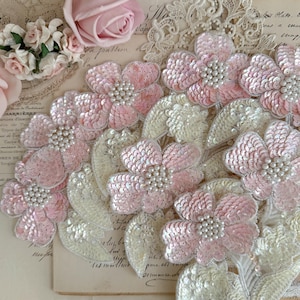 May include: A floral applique featuring light pink sequined flower petals and white sequined leaves. The centre of each flower is embellished with a cluster of small white faux pearls. The design is layered with multiple blossoms and foliage elements.