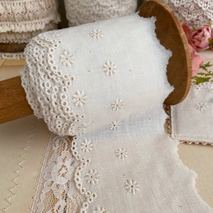 May include: A roll of white fabric with embroidered floral details and a scalloped lace edge. The fabric is wrapped around a wooden spool. Additional rolls of lace are visible in the background, suggesting a craft or sewing theme.