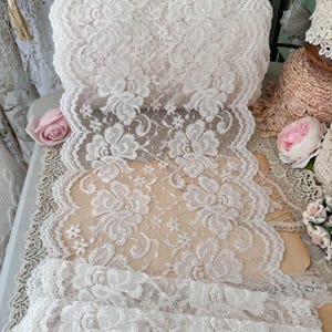 May include: A wide roll of off-white lace fabric featuring a detailed floral pattern with scalloped edges. The delicate textile displays intricate rose motifs woven into a sheer mesh background, showcasing a continuous repeating design throughout.