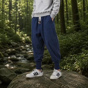 May include: A pair of loose-fitting navy blue trousers with a drawstring waist and fitted cuffs at the ankles, paired with a light gray long-sleeved sweatshirt and white sneakers with black stripes, worn in an outdoor forest setting.