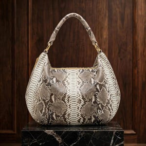 Puede incluir: Un bolso de estilo hobo con un patrón de piel de serpiente en gris y blanco. El bolso tiene un asa arqueada única sujeta con anillos de metal dorado y un cierre de cremallera superior. La superficie texturizada muestra un diseño de escamas.