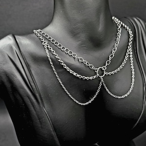 May include: A silver-toned multi-strand chain necklace featuring three distinct layers of metal links connected by a central circular ring. The design includes a thicker top chain and two thinner, draping lower chains that create a layered effect.
