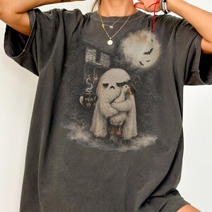 May include: A charcoal gray short-sleeve t-shirt featuring a graphic print of a ghost wearing a sheet and holding a goose that wears a witch hat. The ghost holds a mug with the text MEH and a pumpkin icon. A full moon and bats are in the background.