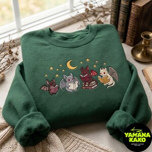 May include: A dark green crewneck sweatshirt featuring an embroidered graphic of four small winged creatures reading books under a yellow crescent moon and stars. The creatures are coloured in shades of brown, grey, dark red, and cream.