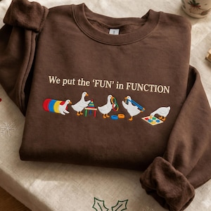 May include: A dark brown crewneck sweatshirt featuring the embroidered text 'We put the 'FUN' in FUNCTION' above a row of five white geese playing with colorful children's toys, including a tunnel, stacking rings, and a shape puzzle.