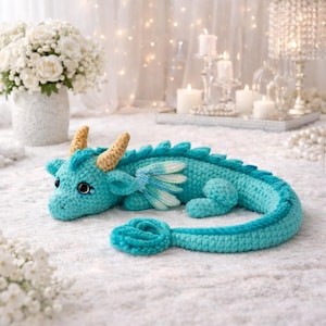 May include: A turquoise crochet dragon plush toy featuring two textured golden horns, small layered wings with white and light blue accents, and a long curled tail. The creature has large dark eyes with blue highlights and a soft yarn texture.