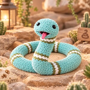 Può includere: Un serpente giocattolo all'uncinetto di colore verde acqua chiaro con strisce orizzontali bianche e verde scuro. Il serpente ha occhi in plastica nera e una lingua in filo rosa brillante che sporge dalla bocca, avvolto su una superficie sabbiosa.