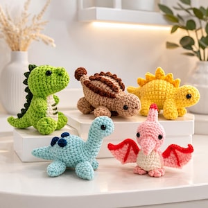 May include: A collection of five small crocheted dinosaur toys in various colours including green, tan, yellow, light blue, and pink. Each plush figure features black bead eyes and distinct textures, with the pink dinosaur displaying red wings.