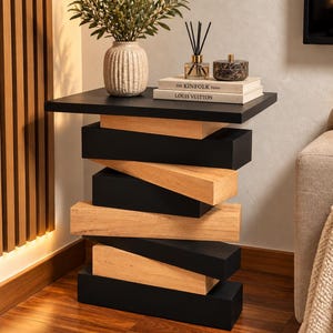 May include: A side table featuring a stacked design of alternating black and light wood rectangular blocks. The table top holds a textured beige vase with greenery, a glass reed diffuser, and two stacked books titled The Kinfolk Home and Louis Vuitton.