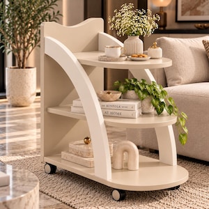 May include: A three-tiered rolling cart in a light beige finish with white curved side supports and four small wheels. The shelves hold a white mug, a small vase with flowers, two books titled The Kinfolk Home and At Home, and decorative objects.