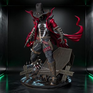 Puede incluir: Una figura detallada de un personaje con sombrero de vaquero negro, capa roja larga y pantalones negros con hebilla de calavera. La figura presenta brazaletes con púas, una pistola y se encuentra sobre una base con un ataúd y dos cruces.