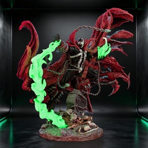 Puede incluir: Una figura de acción detallada de Spawn con traje negro y detalles blancos, una capa roja ondeante y cadenas plateadas. El personaje sostiene efectos de energía verde brillante en ambas manos, sobre una base decorada con calaveras y una lápida.