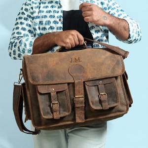 May include: A brown leather messenger bag featuring a top flap with the embossed initials J.M., a central buckle strap, and two smaller front pockets with buckle closures. The bag includes a shoulder strap and metal hardware accents.