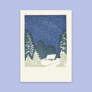 May include: A rectangular art print featuring a winter landscape with a small cabin nestled among snow-covered evergreen trees under a dark blue night sky filled with white stars. The scene is framed by a wide, textured off-white border.