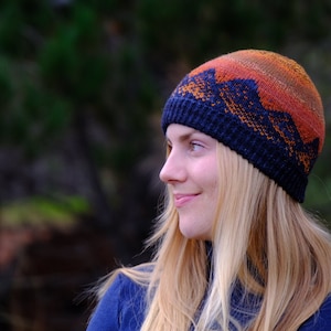 May include: A woman wearing a blue and orange knit hat with a mountain design. The hat is made of a soft, warm yarn and has a comfortable fit.