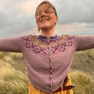 May include: A light purple cardigan with a detailed floral pattern around the neckline and shoulders. The cardigan has a button-up front and long sleeves. The design incorporates shades of purple, yellow, and green. The person is wearing a brown belt and a yellow skirt.