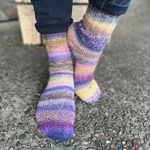 May include: A close-up of two hand-knitted socks. The socks display a striped pattern in shades of purple, yellow, pink, and blue. The socks are made from a soft, textured yarn, suggesting warmth and comfort.