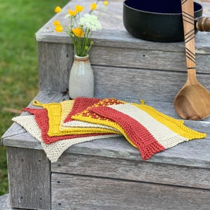 Peut inclure: Plusieurs dessous de plat tricotés aux couleurs vives : rouge, jaune et crème. Ils sont disposés sur une surface en bois vieilli. Un petit vase avec des fleurs jaunes et une cuillère en bois sont également visibles.