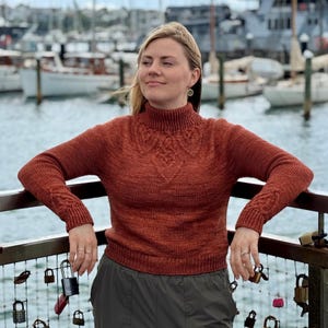 Può includere: Un maglione a collo alto color ruggine con intricati dettagli a trecce sul petto e sulle maniche. Il maglione ha una silhouette aderente ed è realizzato in un filato morbido e strutturato. La modella indossa il maglione all'aperto.