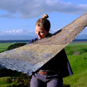 May include: A hand-knitted shawl in shades of blue, yellow, and brown. The shawl has a textured, striped pattern and a scalloped edge. The background features a green landscape and a blue sky.