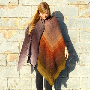 May include: A knitted shawl in a gradient of colors, transitioning from dark purple to shades of brown, orange, and yellow. The shawl is draped over the shoulders, showcasing its textured knit design and color variations. The shawl is made of soft yarn.