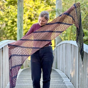 May include: A hand-knitted shawl in brown, purple, and green, with detailed openwork and geometric patterns. The shawl is displayed, highlighting its varied textures and colour combinations. The design includes stripes and diamond shapes.