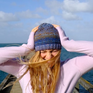 May include: A woman with long blonde hair is wearing a blue and brown knit hat and a pink sweater. Her hair is blowing in the wind and she is holding her head with both hands.