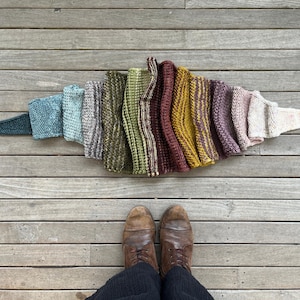 Op de afbeelding: Een verzameling kleurrijke gebreide items, gerangschikt in een waaier, met een spectrum aan tinten van blauwgroen tot crème. De items worden getoond op een houten oppervlak, met een paar bruine leren laarzen op de voorgrond.