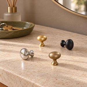 Peut inclure: Quatre boutons de placard aux finitions variées sont disposés sur une surface mouchetée. L'ensemble comprend un bouton en chrome poli, deux boutons en laiton brossé et un bouton noir mat, chacun ayant une forme ronde classique.