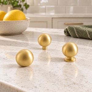 Peut inclure: Trois boutons de placard sphériques avec une finition dorée brossée. Chaque bouton présente une tête ronde et lisse fixée sur une base cylindrique courte, conçue pour être montée sur des portes ou des tiroirs de meubles.