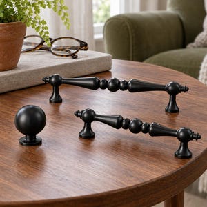 May include: A set of matte black cabinet hardware consisting of two elongated drawer pulls with a central beaded design and one spherical cabinet knob. The hardware pieces are finished in a solid dark tone with a smooth, uniform texture.