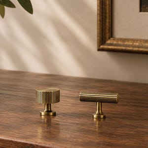 May include: Two brass cabinet knobs with a ribbed, cylindrical texture. One knob is circular and the other is a T-bar shape. Both feature a metallic finish with vertical grooves and a small, smooth pedestal base for mounting to furniture.