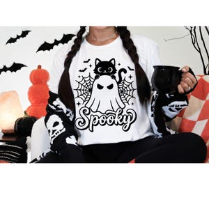 以下が含まれることがあります： 白い長袖Tシャツ。幽霊の上から顔を出す猫、その周りにクモの巣、コウモリ、小さな星が描かれた黒いグラフィックプリントが特徴です。グラフィックの下部には、スタイリッシュな黒い筆記体フォントで「Spooky」という文字がプリントされています。