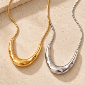 Puede incluir: Dos collares con cadenas de serpiente, uno en tono dorado y otro en tono plateado. Cada collar presenta un colgante curvo en forma de media luna con una superficie texturizada e irregular. Los colgantes están unidos a las cadenas por ambos extremos.
